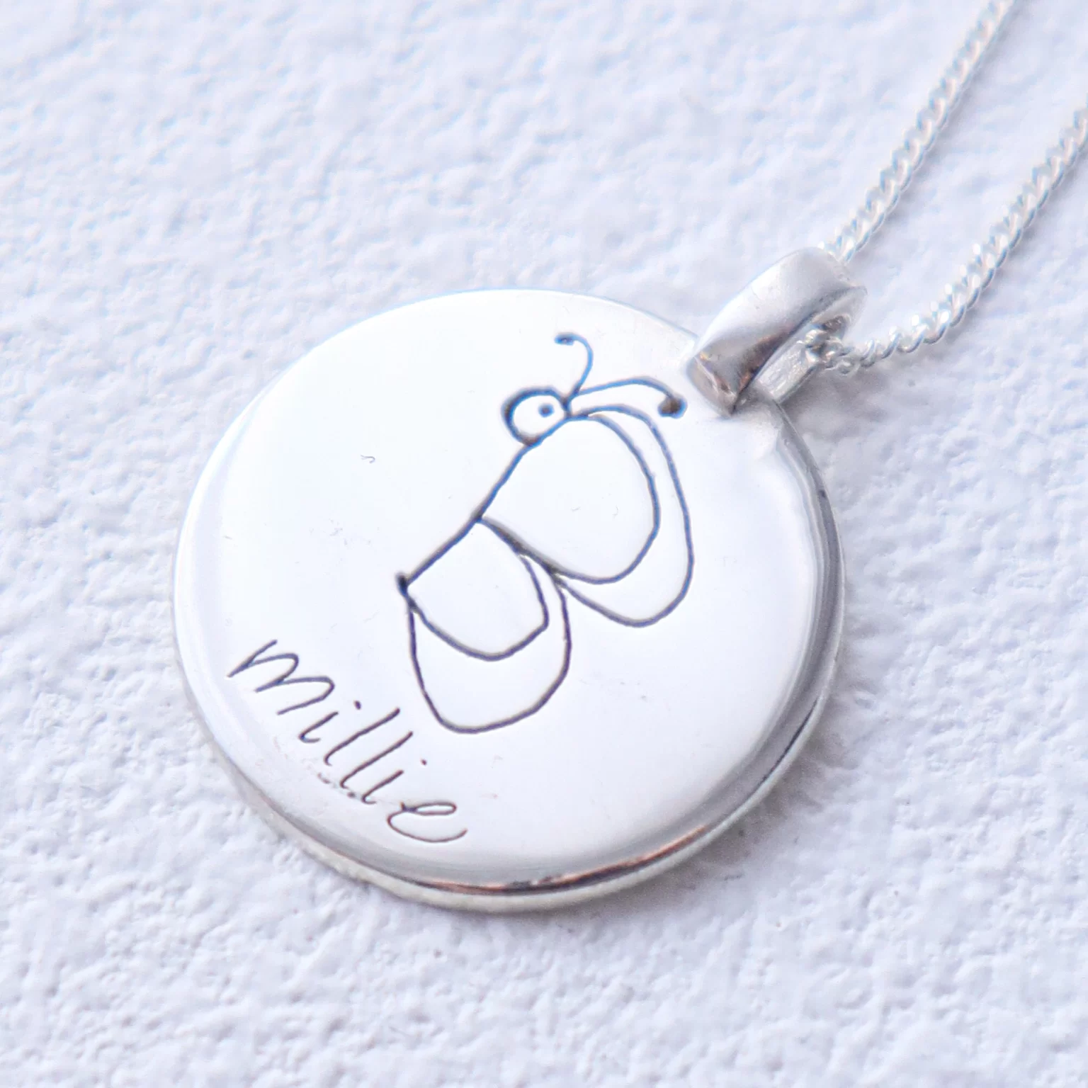 A silver round pendant with a simple line drawing of a butterfly and the name "millie" etched beneath it. This piece of personalised jewellery from Australia hangs elegantly from a thin, silver chain on a textured white surface.