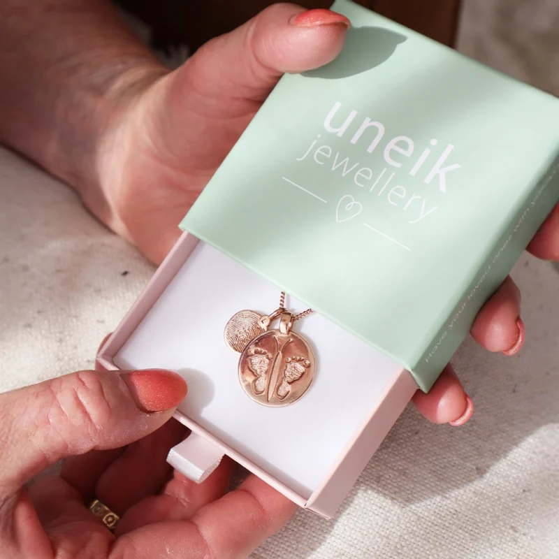 A pair of hands holds a green box with "uneik jewellery" written on it in white letters. Inside the box is a necklace with a circular, intricate pendant alongside an additional small footprint disc pendant, crafted beautifully in Australia.