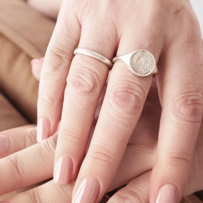 Close-up of two hands with light pink manicured nails, showing rings on both. One ring is a simple, thin band, and the other is a larger signet ring with a fingerprint design. Hands are resting on a light brown surface.